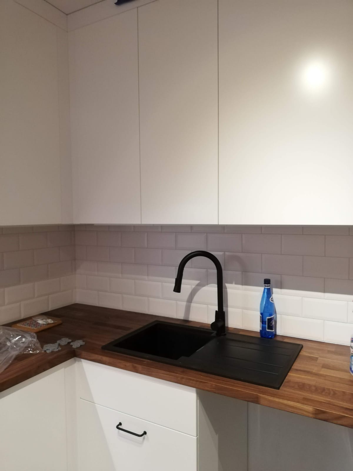 Nowoczesna kuchnia z białymi szafkami, drewnianym blatem i czarnym zlewozmywakiem z baterią; na blacie stoi butelka wody mineralnej.