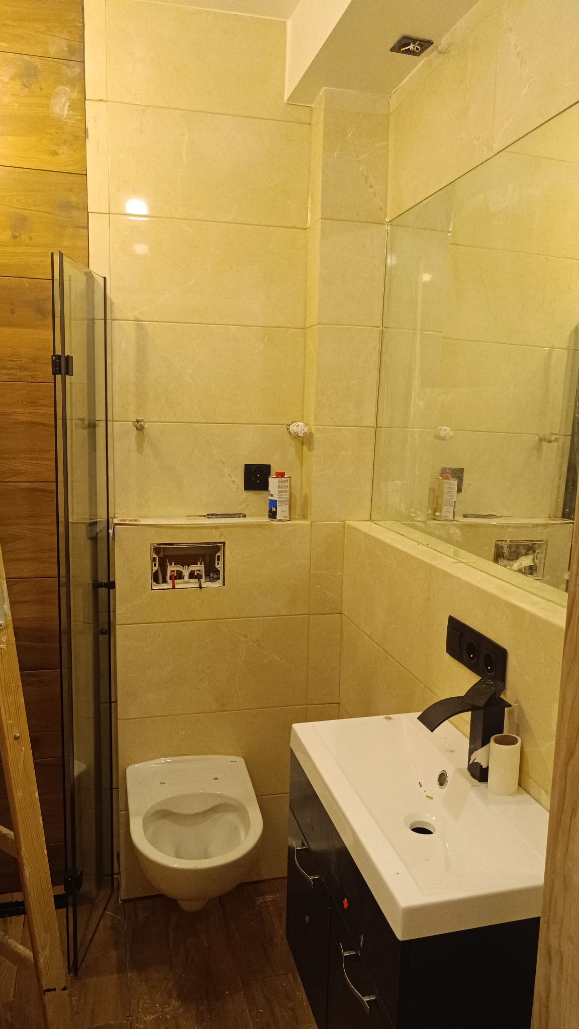 Wnętrze łazienki w trakcie montażu: podwieszana toaleta, umywalka z czarną armaturą, płytki w odcieniach beżu, widoczne elementy instalacji i drabina malarska.