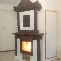 Kaflowy kominek z ciemnobrązową, rzeźbioną obudową i paleniskiem z widocznym ogniem, na tle białej ściany i kafelkowej podłogi.