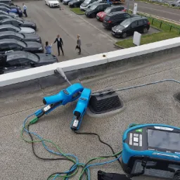 Urządzenie pomiarowe Metrel EurotestXD MI 3155 z niebieskimi klemami i przewodami na szarym dachu, w tle parking z samochodami i ludźmi.