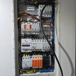 Otwarta skrzynka elektryczna z zainstalowanymi bezpiecznikami, licznikiem i okablowaniem w kolorach niebieskim, brązowym i żółto-zielonym. Widoczne oznaczenia na elementach i żółte oznaczniki...