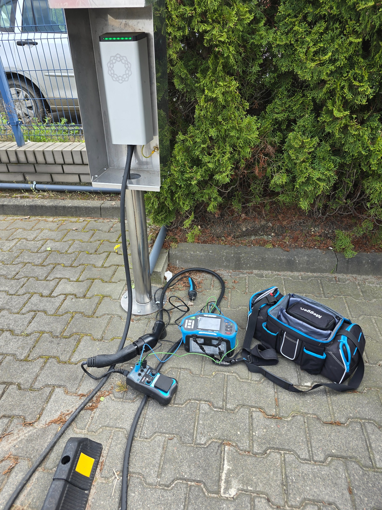 Stanowisko ładowania pojazdów elektrycznych z podłączonym sprzętem pomiarowym Megger i torbą narzędziową na brukowanym chodniku, z krzewem w tle.