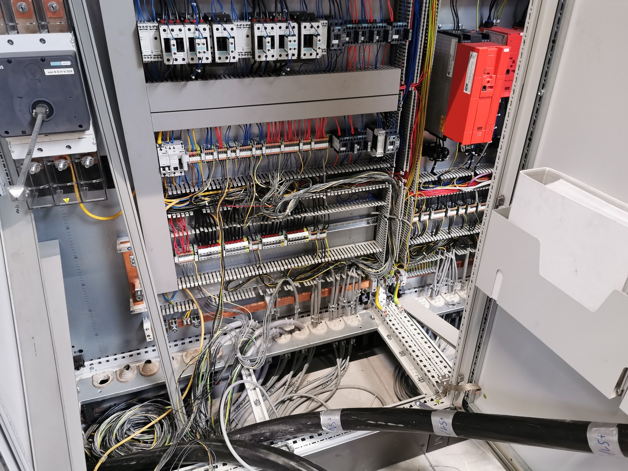 Otwarta szafa sterownicza z widocznymi przewodami, bezpiecznikami, stycznikami i modułami sterującymi SEW, z czarnymi kablami wychodzącymi z dołu szafy.