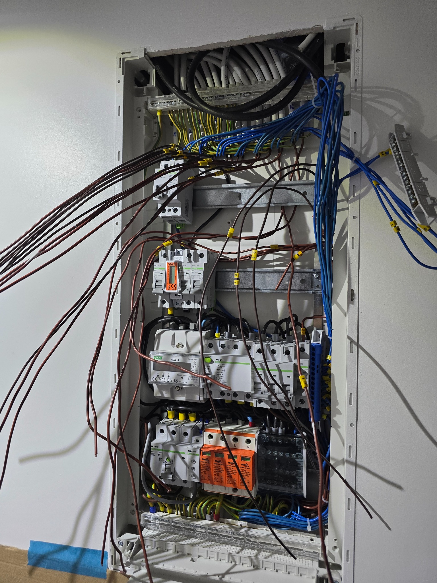 Otwarta skrzynka elektryczna z widocznymi przewodami o różnych kolorach (brązowe, niebieskie, żółto-zielone) i elementami elektrycznymi, takimi jak bezpieczniki i liczniki, z żółtymi oznaczeniami...