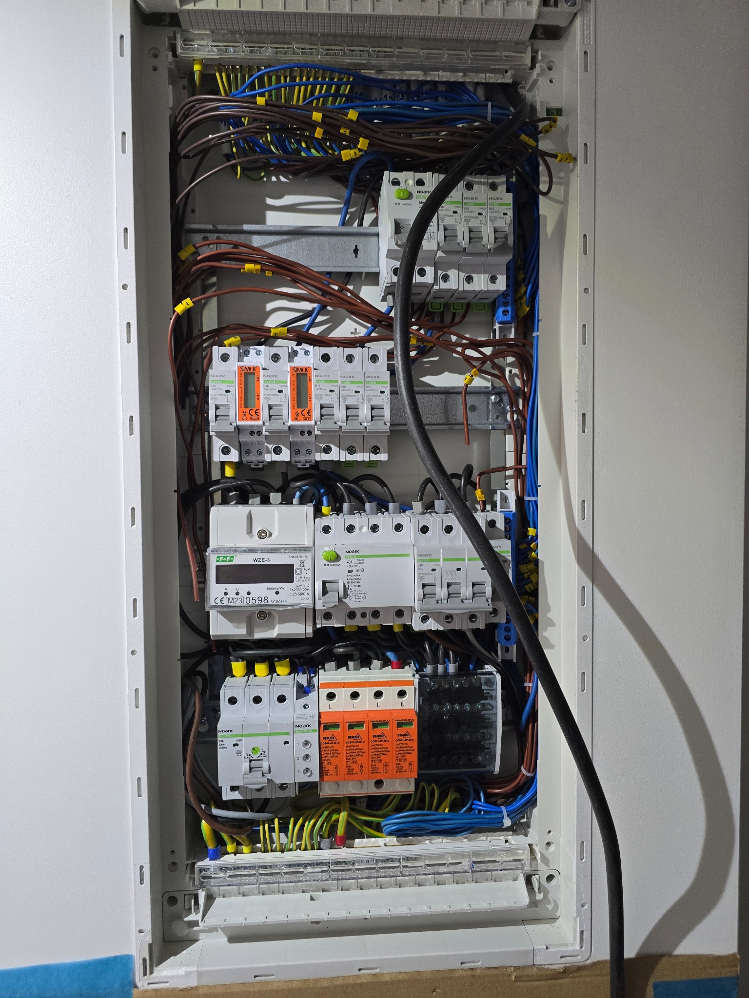 Otwarta skrzynka elektryczna z zainstalowanymi bezpiecznikami, licznikiem i okablowaniem w kolorach niebieskim, brązowym i żółto-zielonym. Widoczne oznaczenia na elementach i żółte oznaczniki...