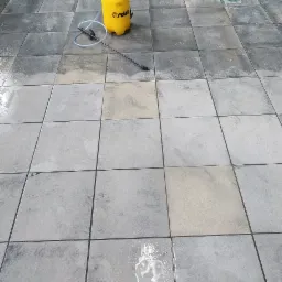 Szare płytki chodnikowe częściowo umyte, z widocznym żółtym opryskiwaczem ciśnieniowym i lancą.