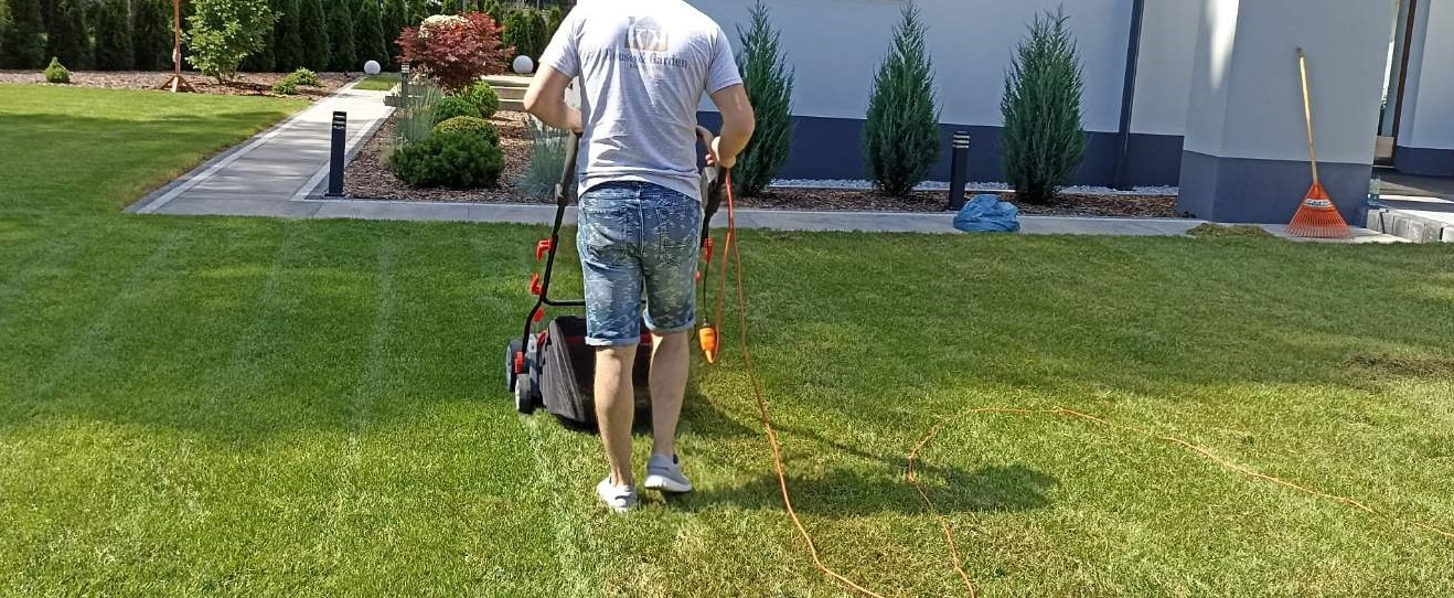 Mężczyzna od tyłu kosiarką elektryczną, widoczny kabel zasilający, równo przystrzyżony trawnik przed nowoczesnym domem z tujami i ozdobnymi kamieniami.