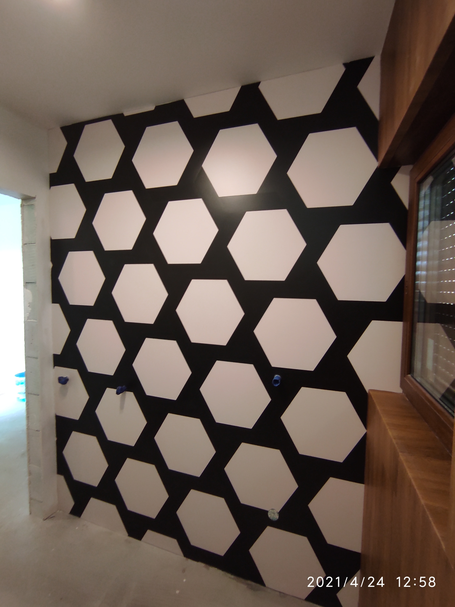 Ściana z geometrycznym wzorem w biało-czarne heksagony, widoczne gniazdka elektryczne i fragment okna z drewnianą ramą. Wnętrze w trakcie wykończenia.