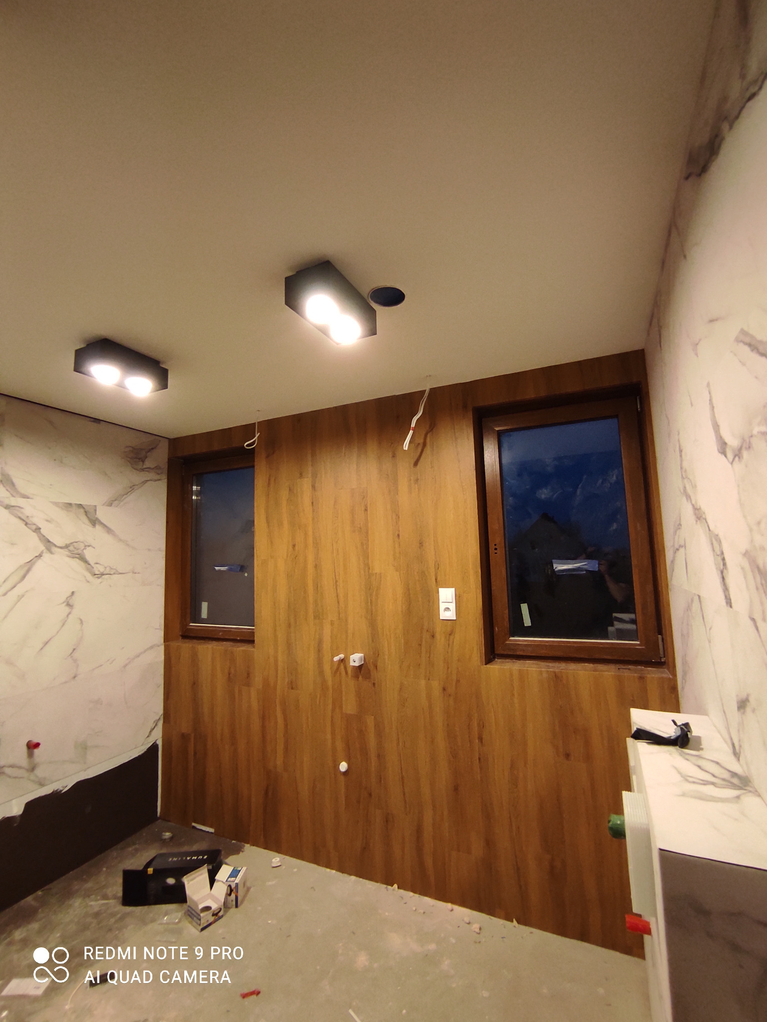 Wnętrze łazienki w trakcie remontu: ściana wyłożona panelami imitującymi drewno, marmurowe płytki, widoczne okna, instalacja elektryczna i sanitarne, narzędzia na podłodze.