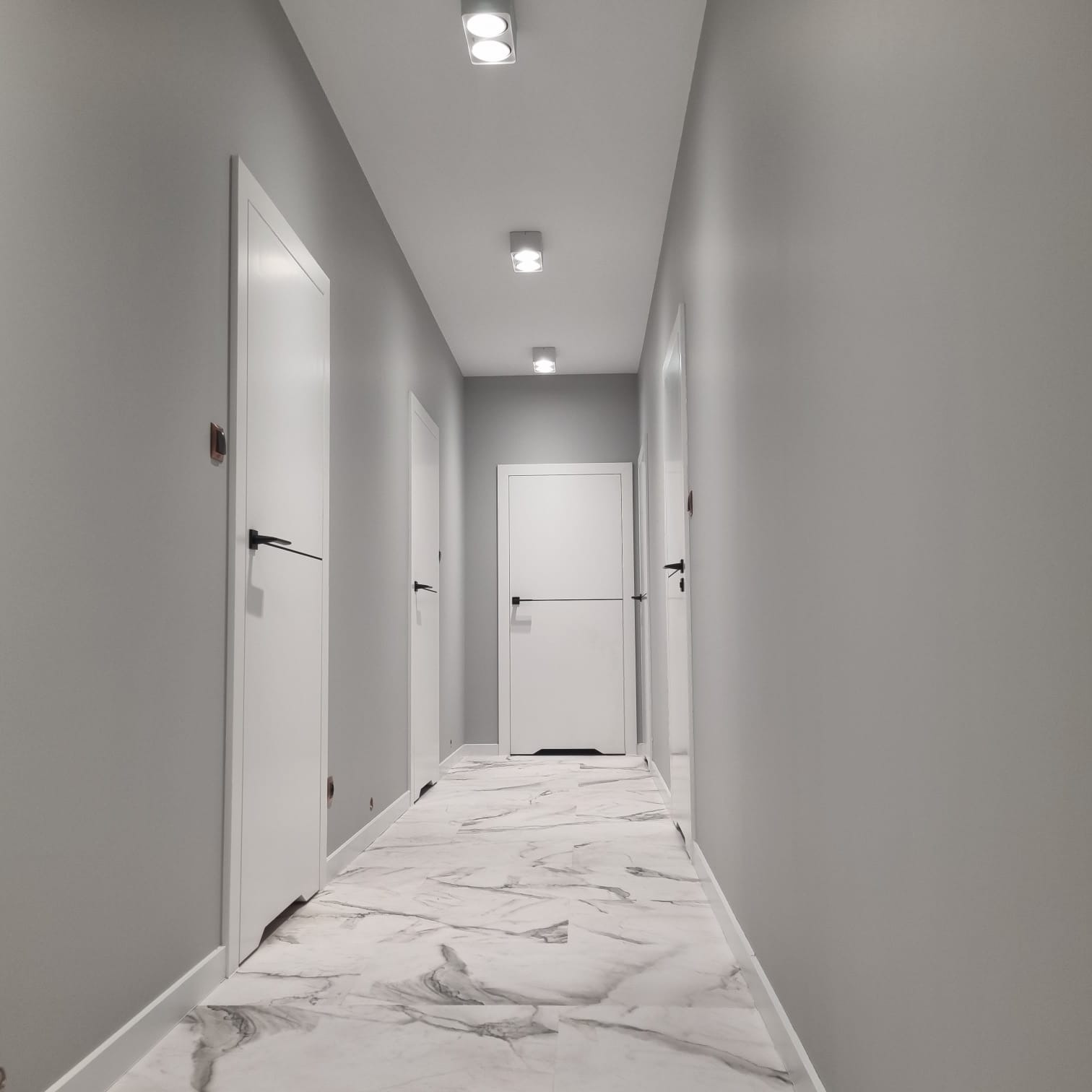 Korytarz w odcieniach szarości z podłogą imitującą biały marmur, białe drzwi z czarnymi klamkami i listwy przypodłogowe, oświetlenie sufitowe punktowe.