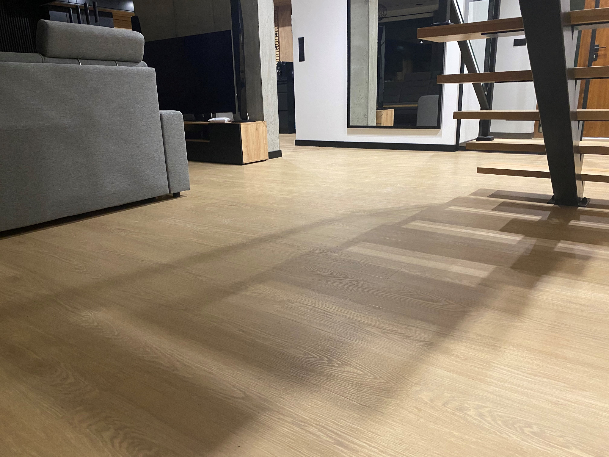 Jasne panele podłogowe w salonie z szarą sofą i schodami z metalową konstrukcją, widoczne cienie stopni na podłodze.