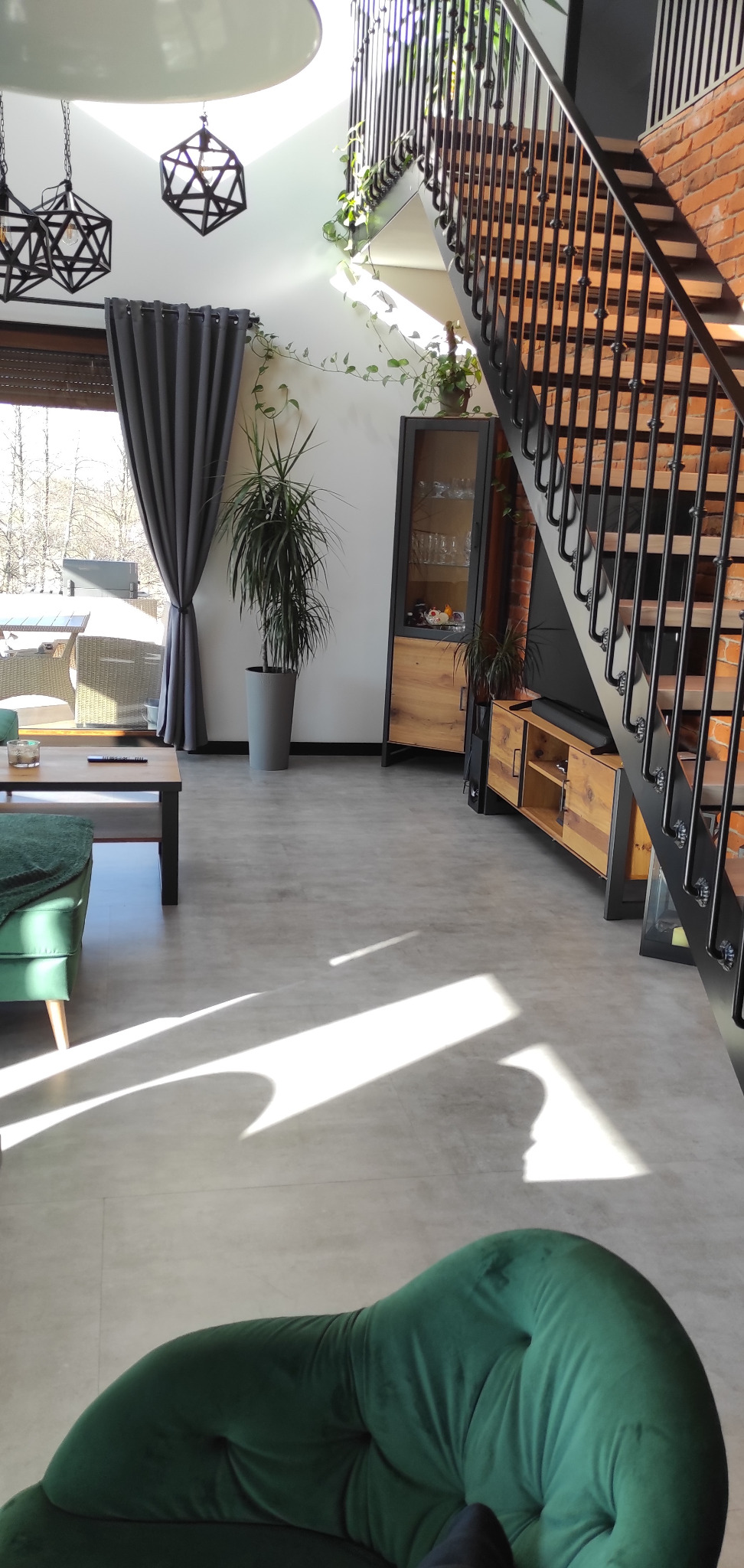 Jasnoszara posadzka w salonie z zieloną sofą, schodami z metalową balustradą i ceglaną ścianą, doświetlona promieniami słonecznymi.