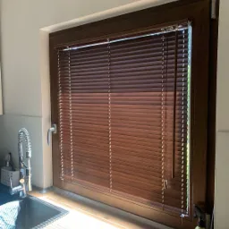 Brązowe poziome listewki w oknie kuchni, z widocznym fragmentem zlewu i blatu, oraz butelką ze spryskiwaczem.