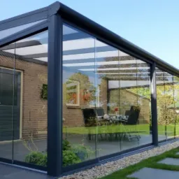 Szklana zabudowa tarasu z metalową konstrukcją w kolorze grafitowym, widok z zewnątrz na stół i krzesła wewnątrz, w tle ceglany budynek i zadbany ogród z kamienną ścieżką.