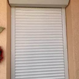 Zamknięta, biała osłona okienna zewnętrzna z widoczną kasetą i prowadnicami na ścianie o fakturze baranka.