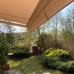 Beżowa osłona przeciwsłoneczna nad tarasem, widok na ogród z zielenią i dom w tle, częściowo rozpakowane elementy montażowe na pierwszym planie.