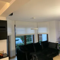 Nowoczesne, białe tekstylne osłony okienne w salonie z czarną sofą narożną, widok na samochód za oknem.