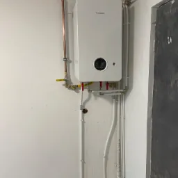 Biały piec gazowy Junkers zamontowany na ścianie z widocznymi rurami miedzianymi i plastikowymi.