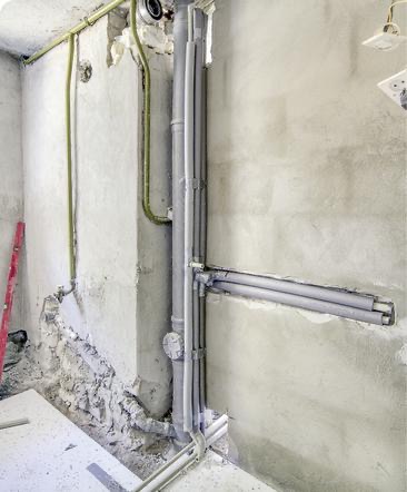 Surowe wnętrze w trakcie remontu: szare rury grzewcze biegnące wzdłuż ściany, fragment instalacji elektrycznej z zielonymi kablami, widoczny fragment różowej poziomicy.