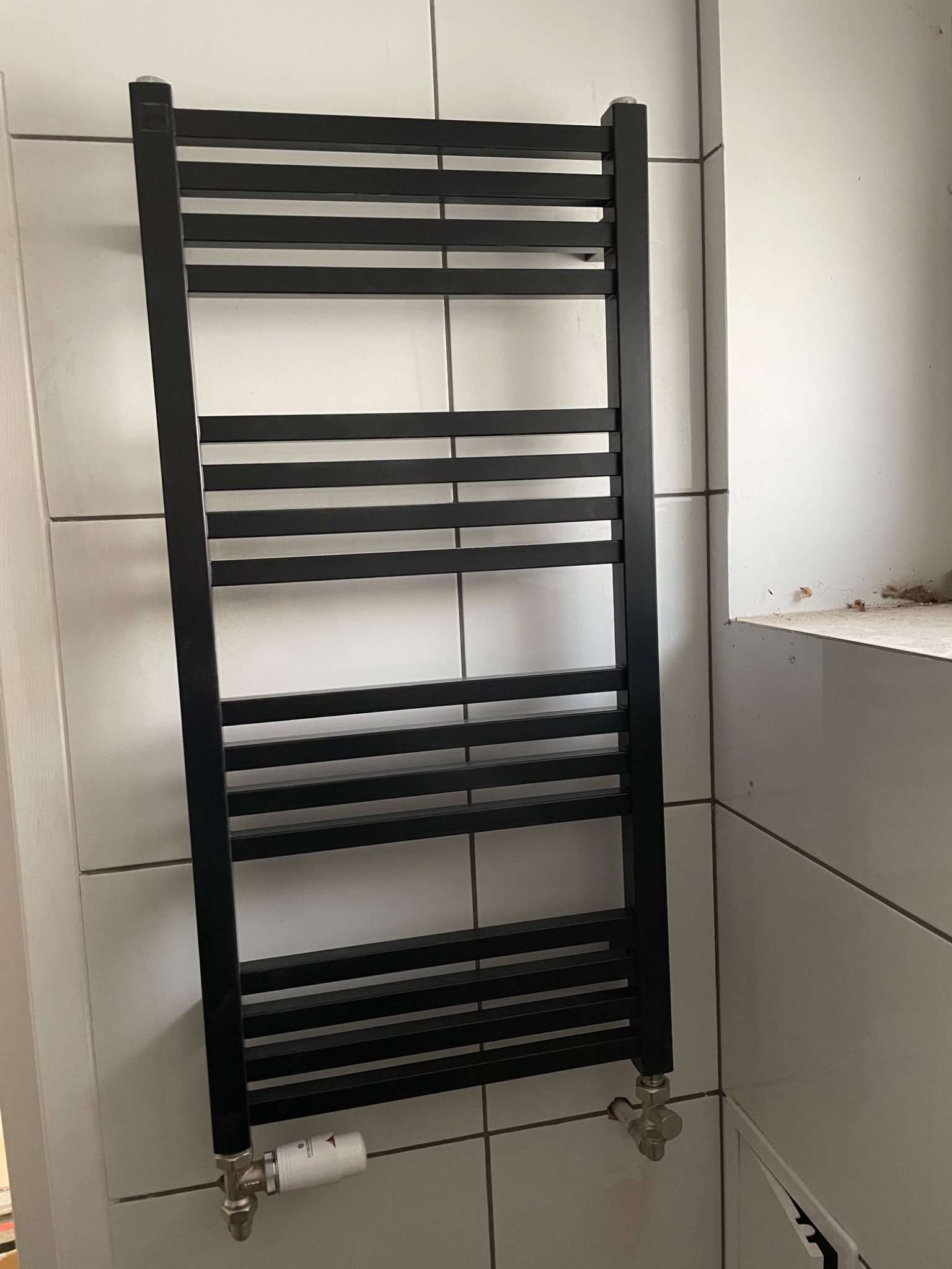 Czarny, drabinkowy grzejnik łazienkowy zamontowany na białej, kafelkowej ścianie, widoczne podłączenie z termostatem.