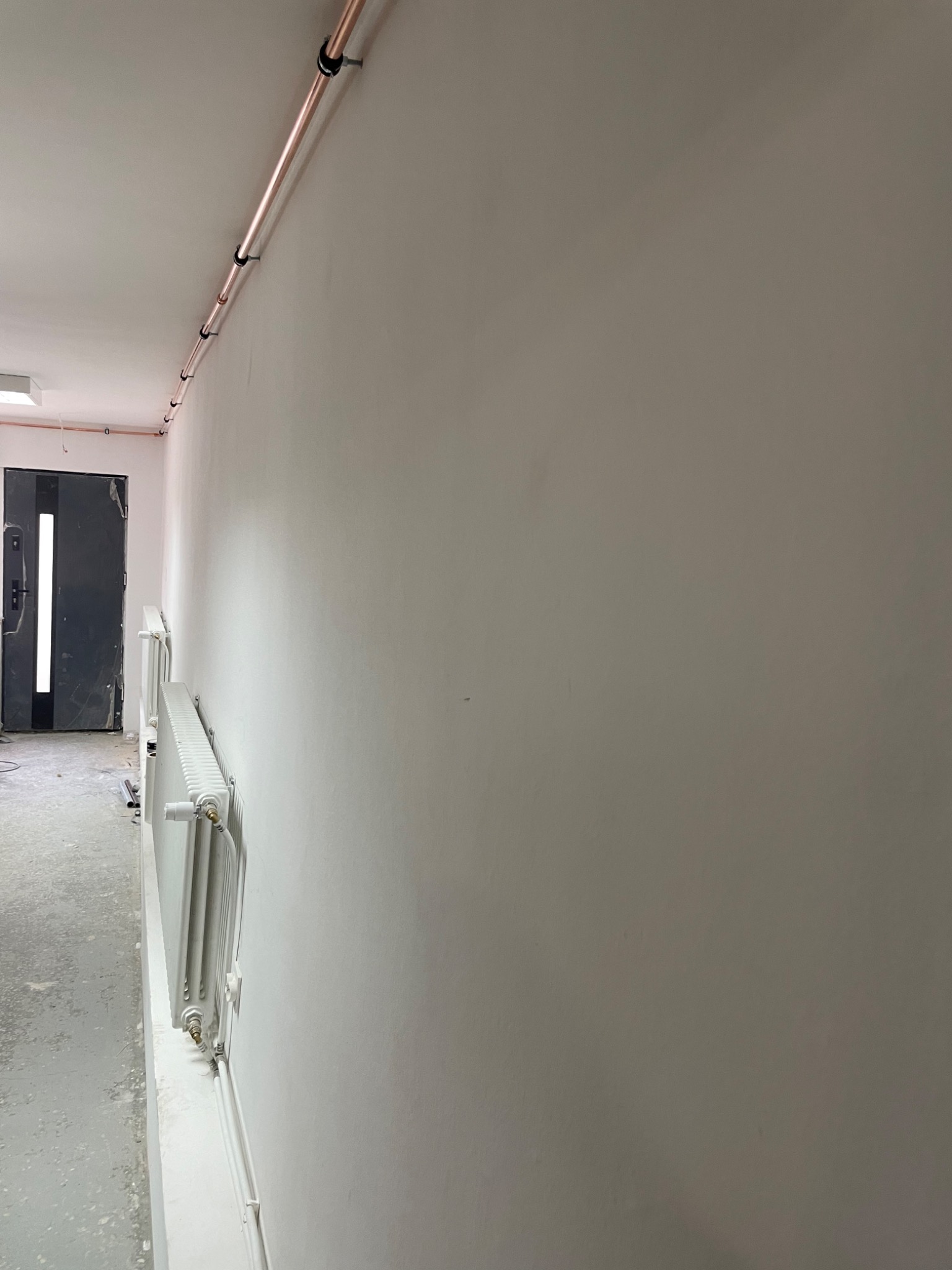 Nowe miedziane rury grzewcze zamocowane na ścianie pod sufitem, z białymi grzejnikami zamontowanymi poniżej, w trakcie remontu korytarza.