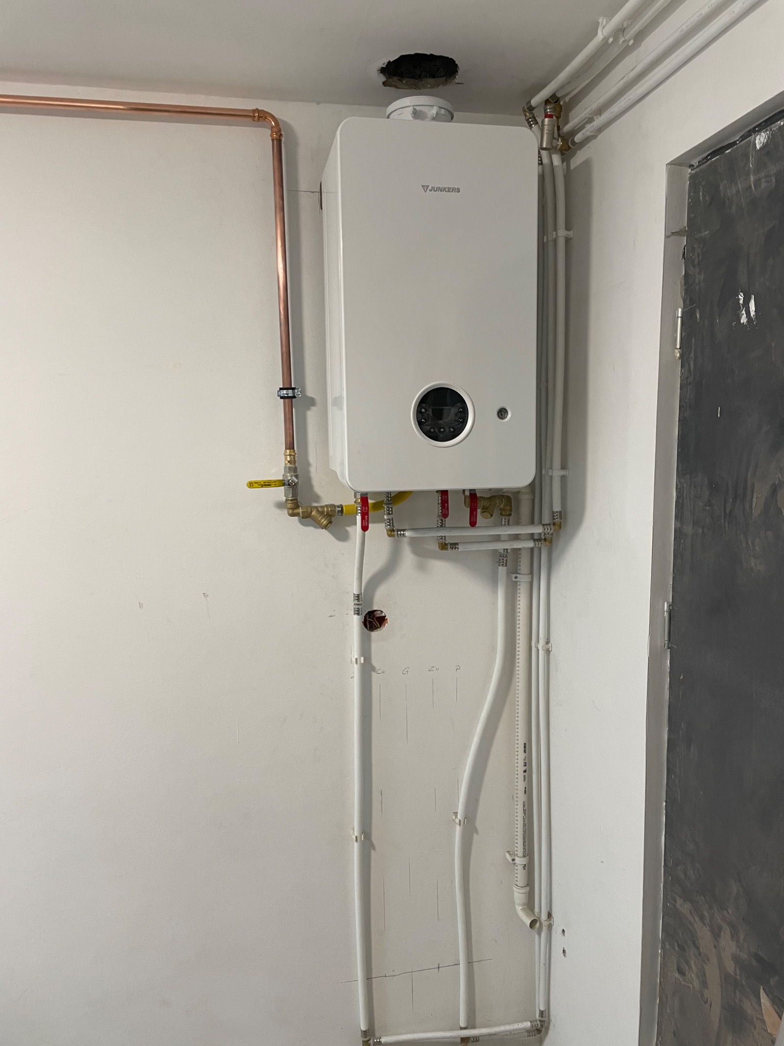 Biały piec gazowy Junkers zamontowany na ścianie z widocznymi rurami miedzianymi i plastikowymi.