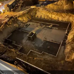 Wykop pod fundamenty zalany betonem, widoczna stal zbrojeniowa, oświetlenie nocne, teren budowy zabezpieczony ziemią.