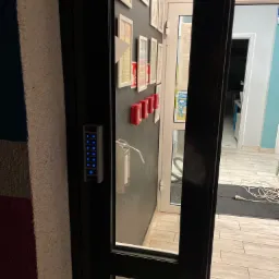 Szare drzwi z zamontowanym panelem numerycznym z podświetlanymi na niebiesko cyframi, widok na wnętrze biura z podłogą wyłożoną płytkami imitującymi drewno.