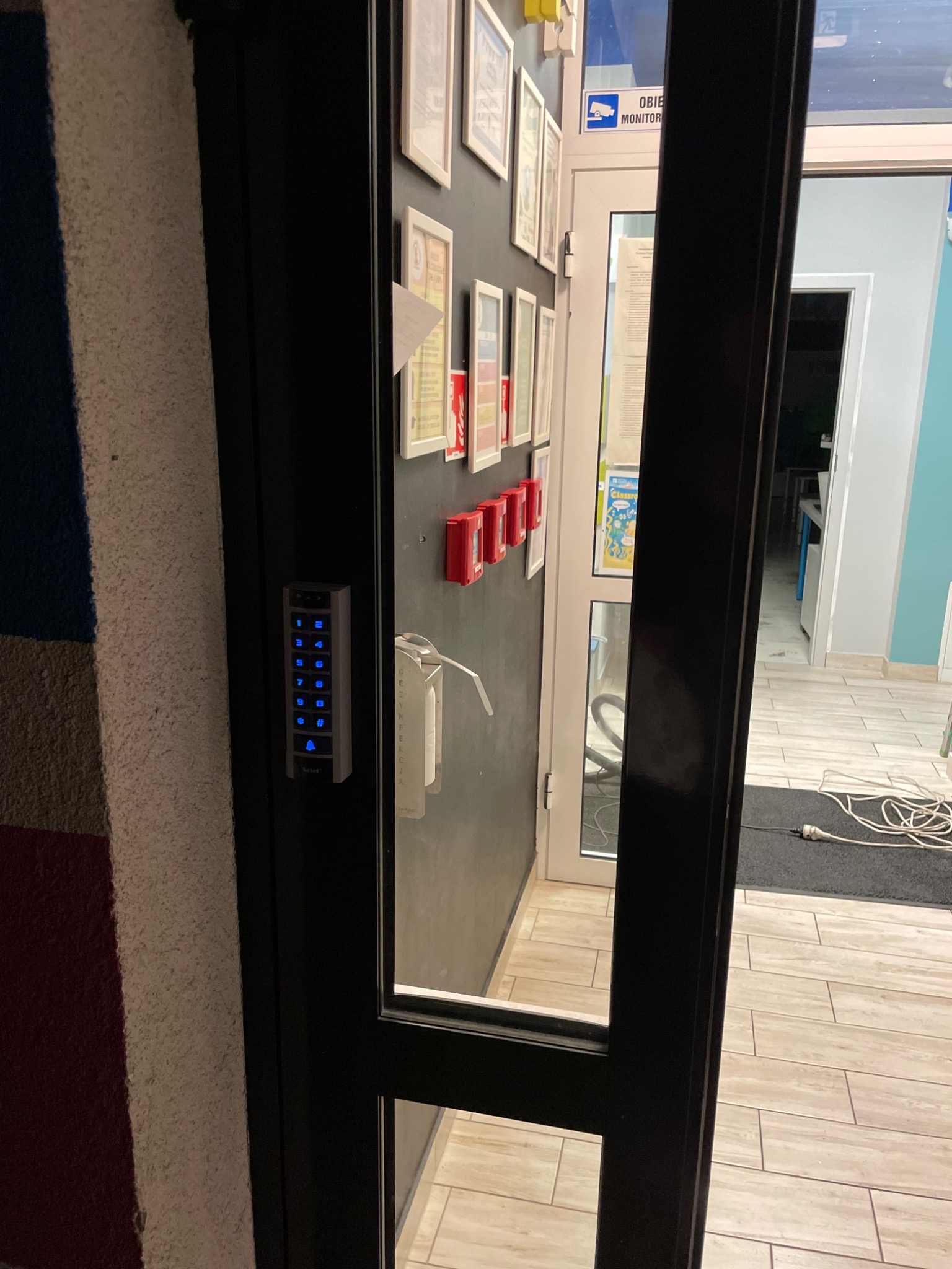 Szare drzwi z zamontowanym panelem numerycznym z podświetlanymi na niebiesko cyframi, widok na wnętrze biura z podłogą wyłożoną płytkami imitującymi drewno.