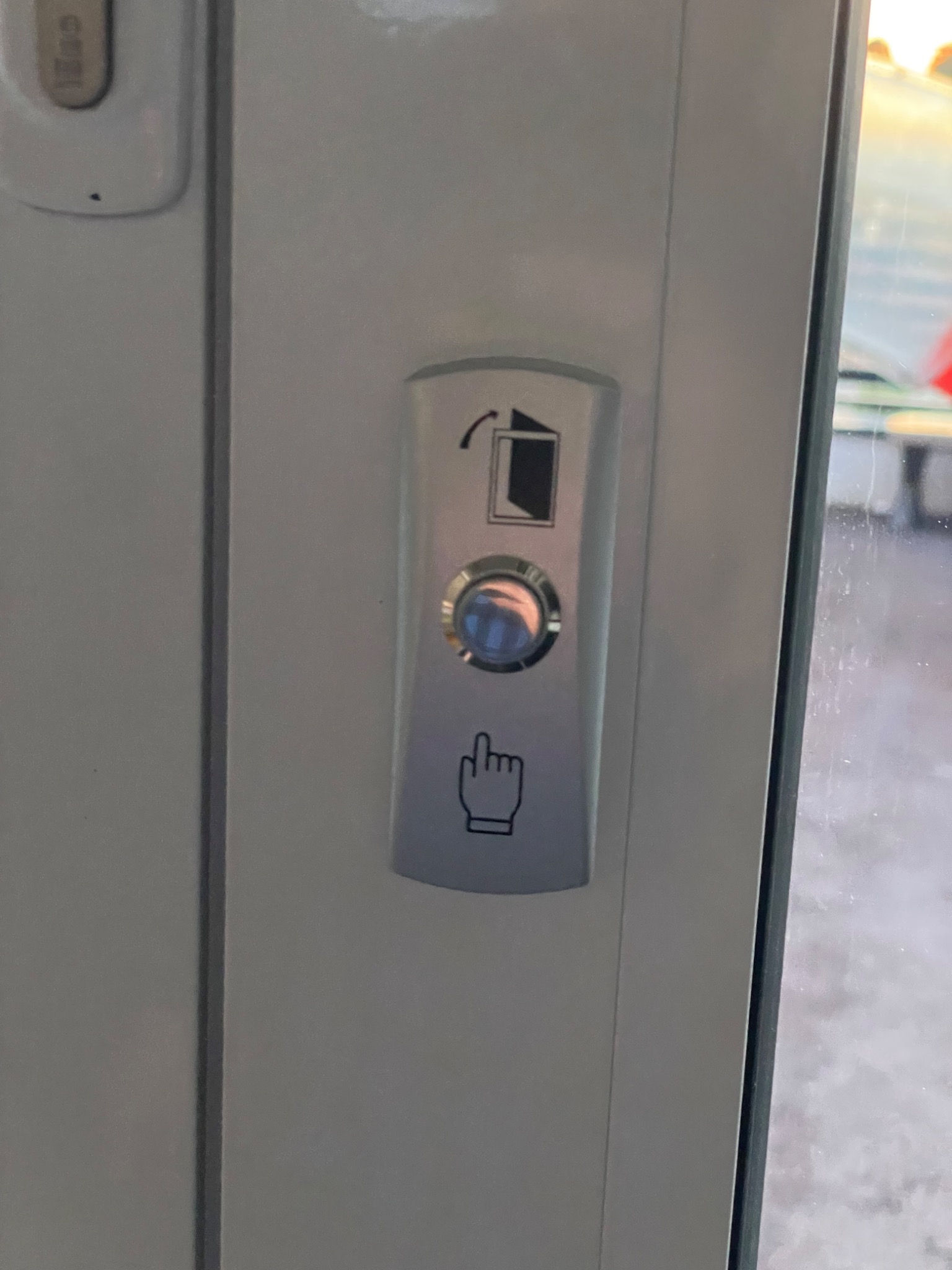 Przycisk otwierania drzwi z symbolem drzwi i dłoni wskazującej, zamontowany na białej ramie drzwi.