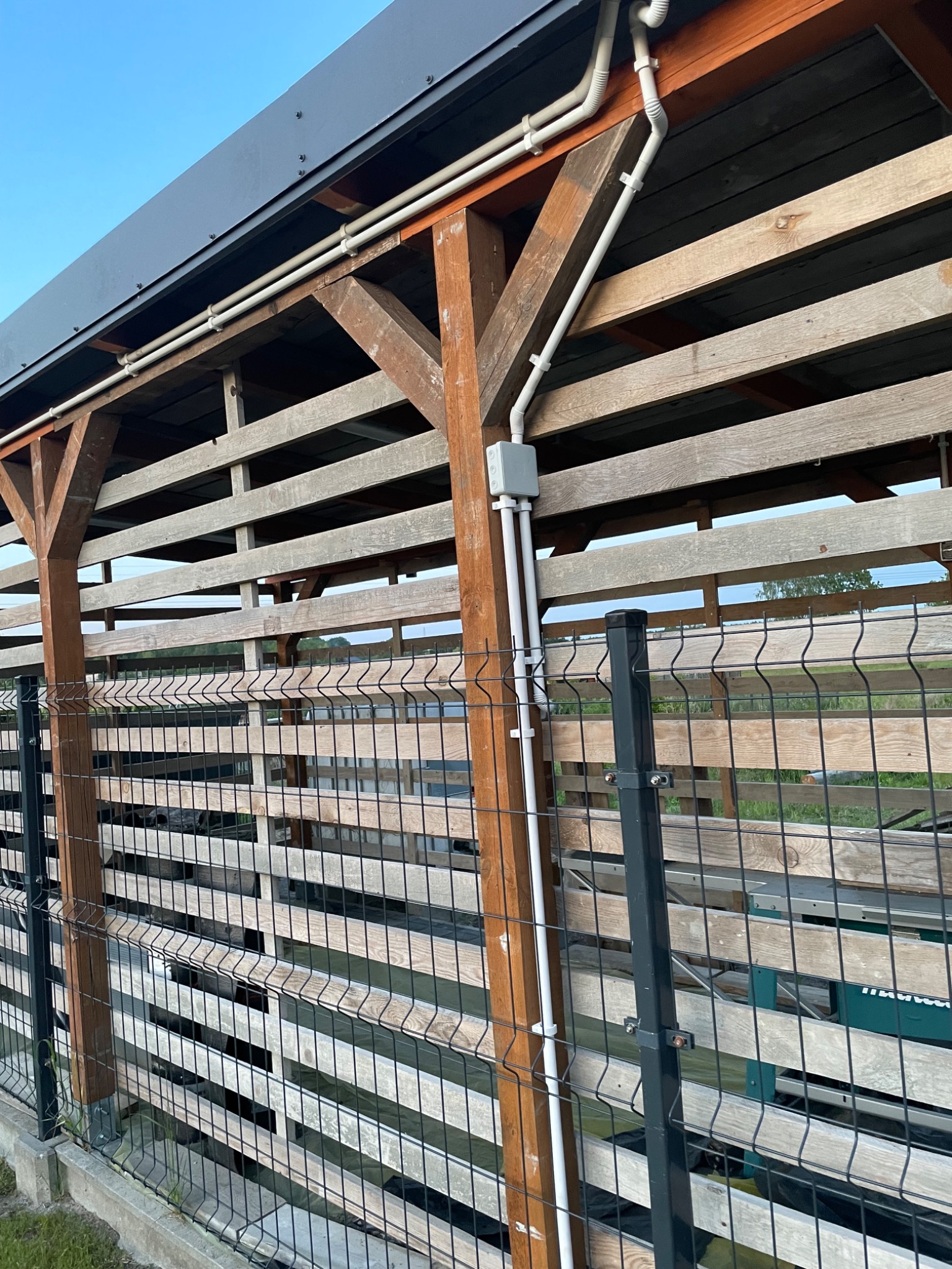 Białe rury elektryczne poprowadzone na zewnątrz drewnianej konstrukcji z metalową siatką, widoczne puszki instalacyjne.