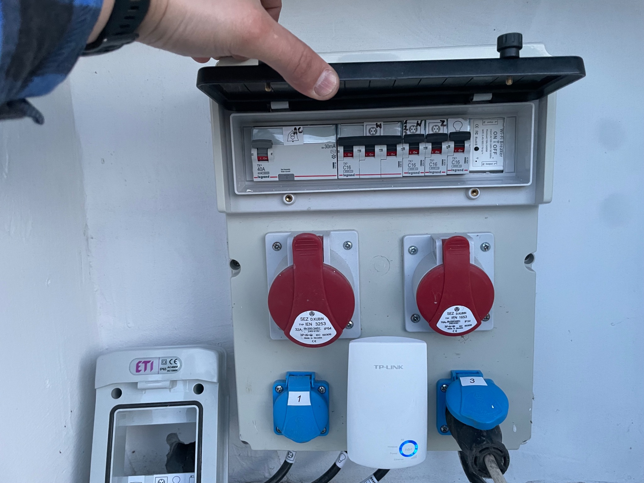 Otwarta skrzynka elektryczna z bezpiecznikami, gniazdami przemysłowymi i modułem Wi-Fi, zamontowana na białej ścianie, z widoczną dłonią otwierającą pokrywę.