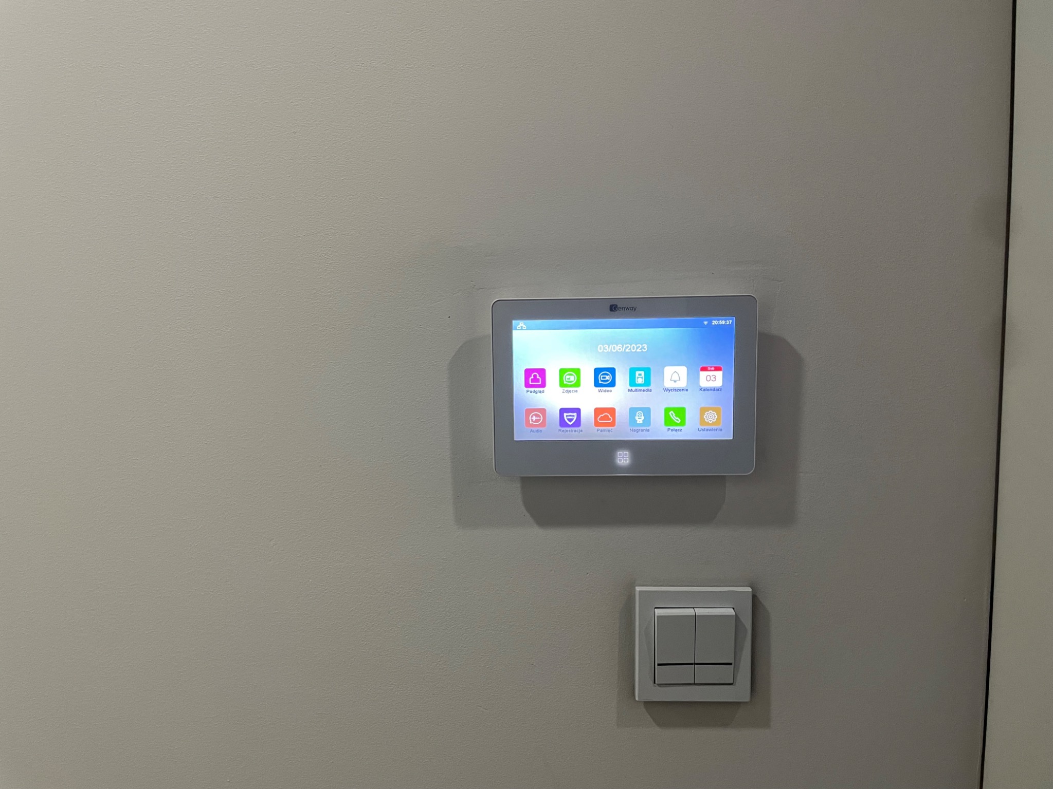 Panel dotykowy wideodomofonu marki Genway zamontowany na ścianie, widoczny obok włącznik światła, ekran wyświetla menu z ikonami i datą 03/06/2023.