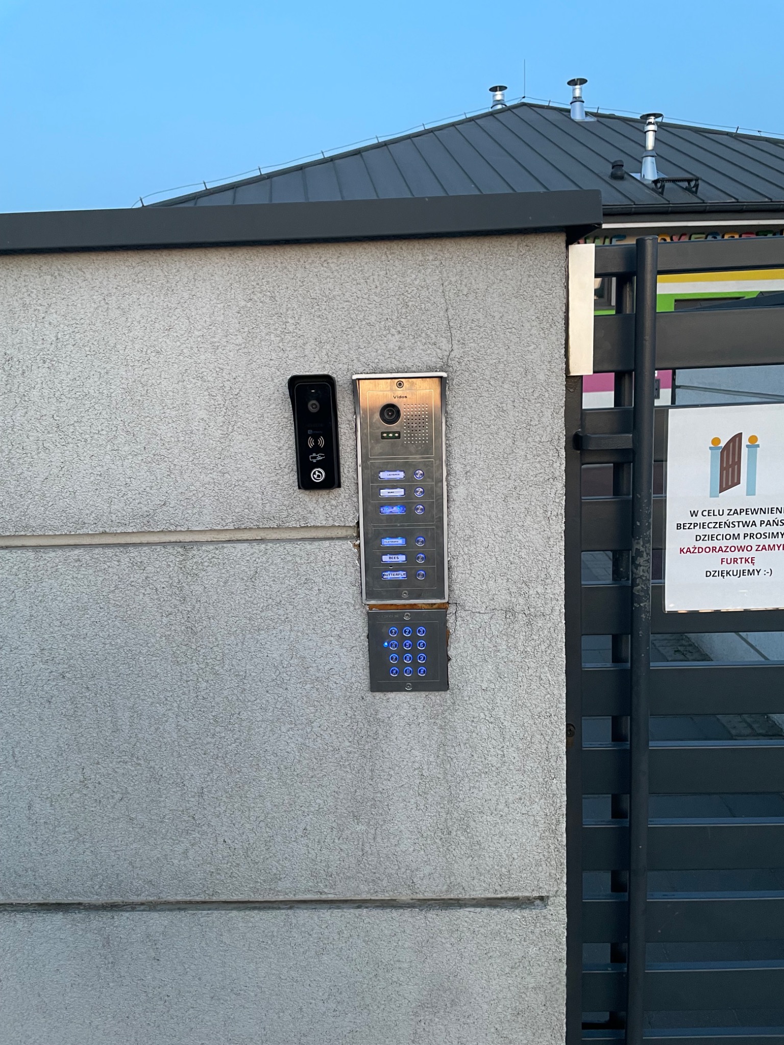 Zestaw domofonowy z kamerą i panelem numerycznym, zamontowany na szarej ścianie obok czarnej furtki z tabliczką informacyjną. Widoczne elementy systemu kontroli dostępu.
