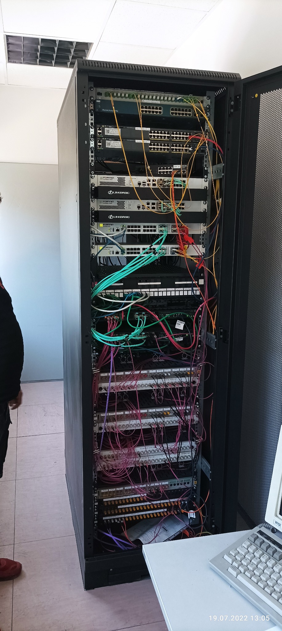 Szafa rack z zainstalowanymi urządzeniami sieciowymi i chaotycznym okablowaniem w kolorach żółtym, zielonym, różowym i fioletowym, widoczne switche Fortinet i Linkbasic, obok biurko z klawiaturą.