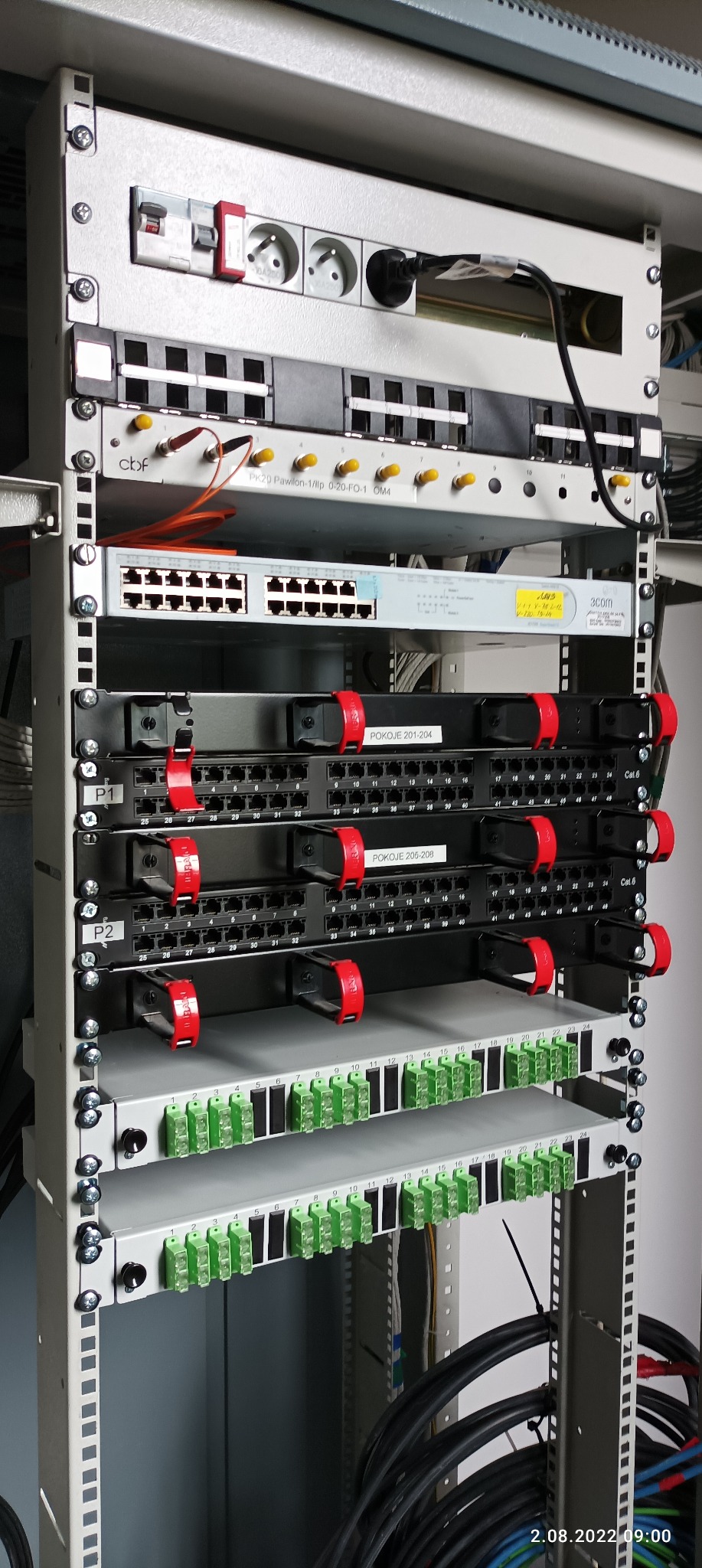 Szczegółowe ujęcie szafy rack z patch panelem światłowodowym, przełącznikiem sieciowym 3com i listwami zaciskowymi, z widocznymi kablami i gniazdami zasilającymi.