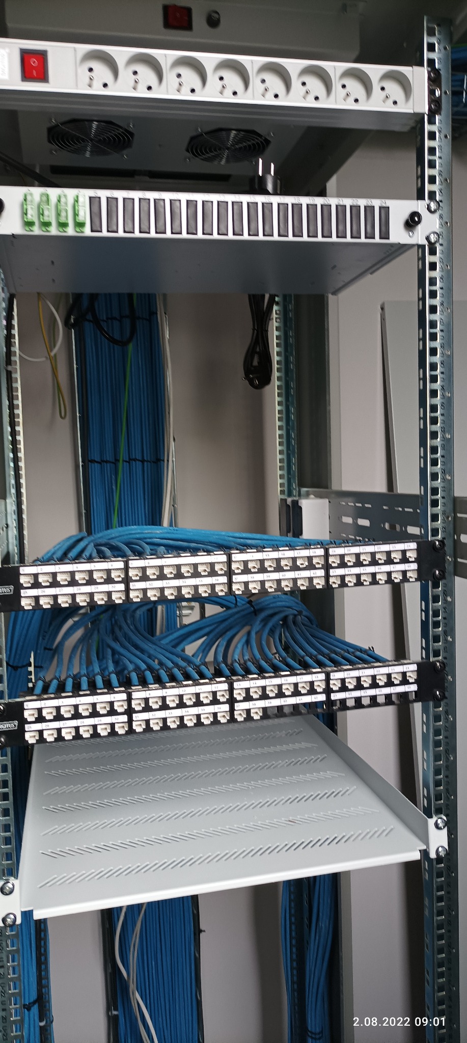 Panel krosowniczy z podłączonymi niebieskimi kablami sieciowymi w szafie rack, widoczne gniazda z numeracją. Nad panelem listwa zasilająca z gniazdami i wentylatorami.