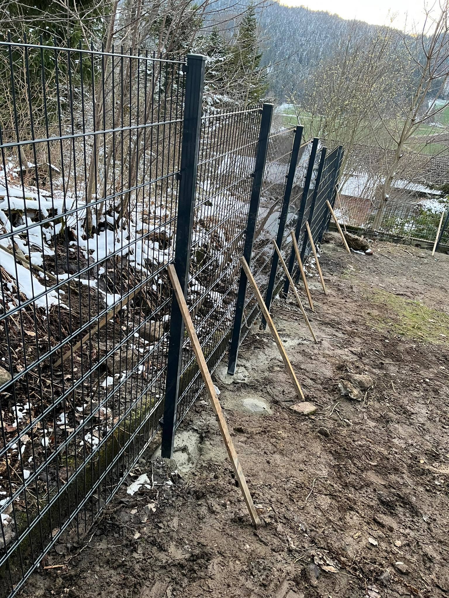 Montaż czarnego ogrodzenia panelowego w terenie z resztkami śniegu, podpory z drewnianych łat stabilizują słupki w świeżym betonie.