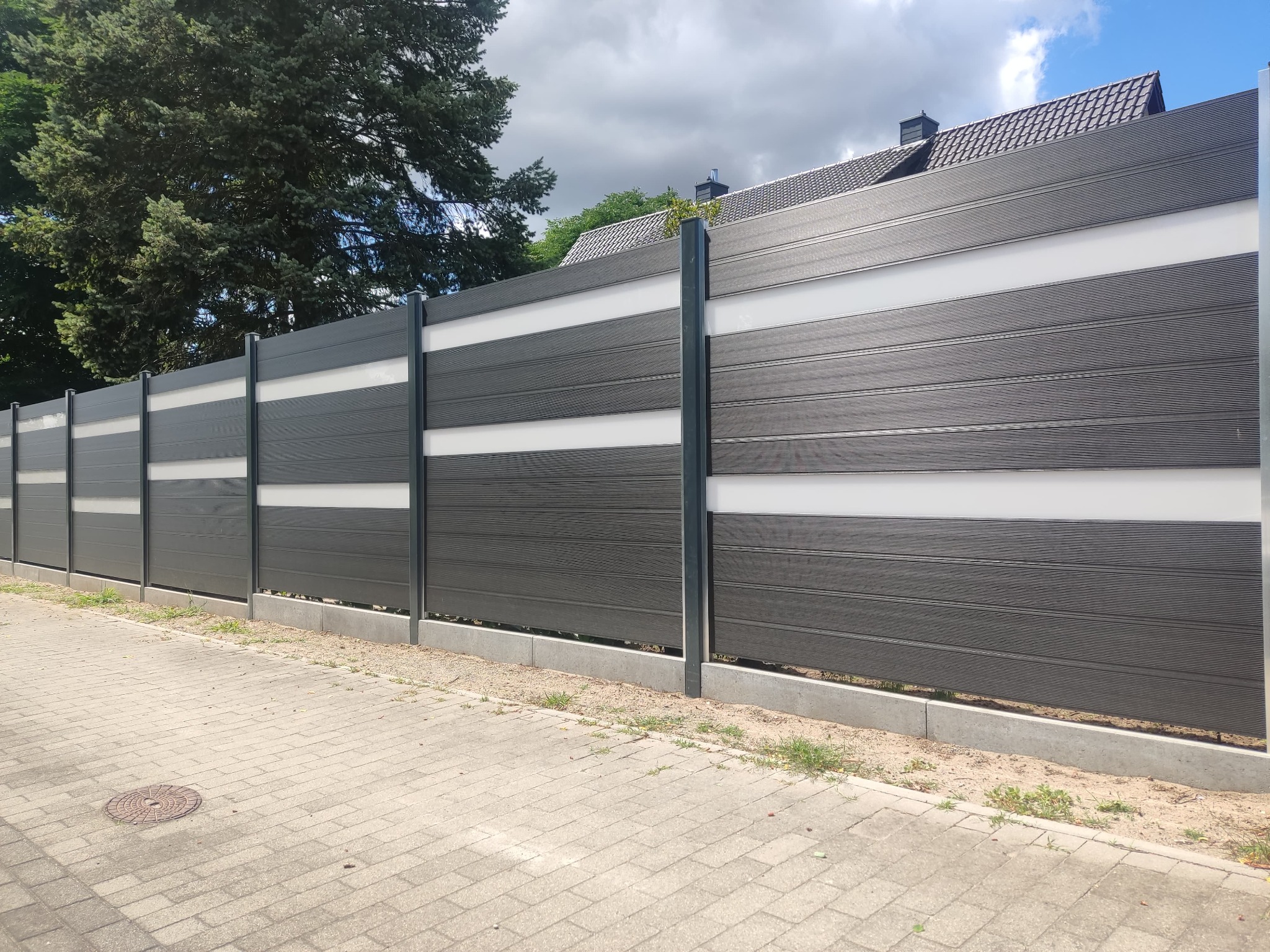 Nowoczesne ogrodzenie panelowe z poziomymi, ciemnoszarymi deskami i białymi wstawkami, wsparte na metalowych słupkach, widoczne z poziomu chodnika z betonowej kostki.