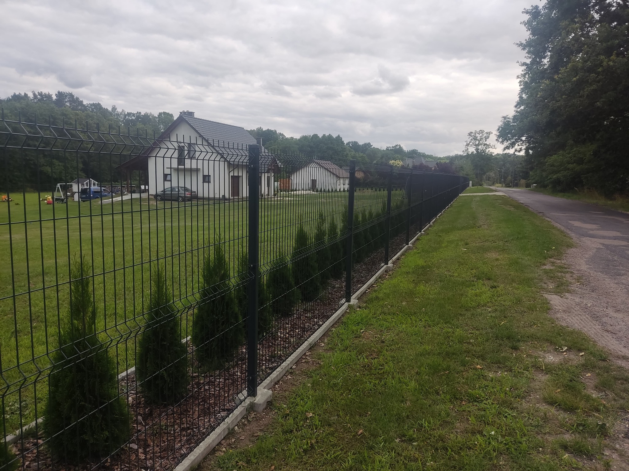 Długa, czarna siatka ogrodzeniowa z podmurówką i nasadzonymi tujami, oddzielająca posesję z białymi domami od drogi asfaltowej i trawnika w pochmurny dzień.