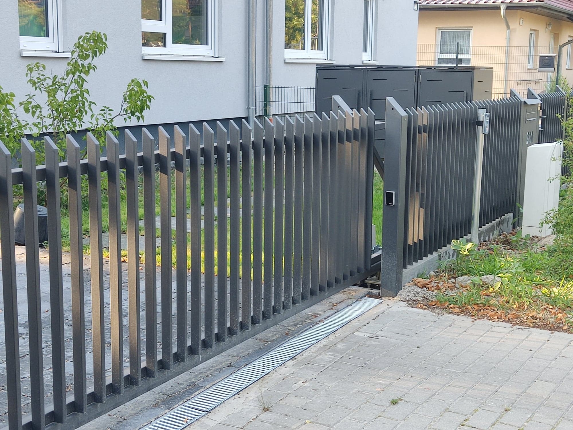 Nowoczesna, przesuwna brama w kolorze antracytowym z pionowymi profilami, widoczny fragment odwodnienia liniowego na kostce brukowej, dom w tle.
