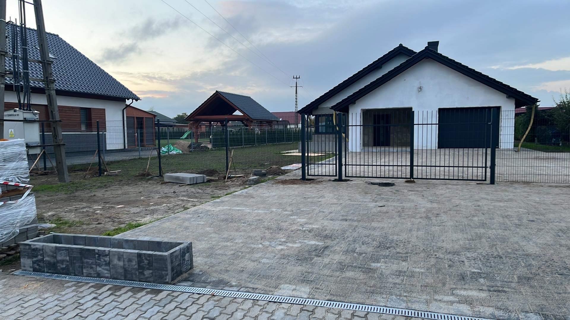 Nowoczesna posesja z szarą kostką brukową, metalową bramą wjazdową i ogrodzeniem panelowym w trakcie montażu, widoczne słupki ogrodzeniowe i materiały budowlane.