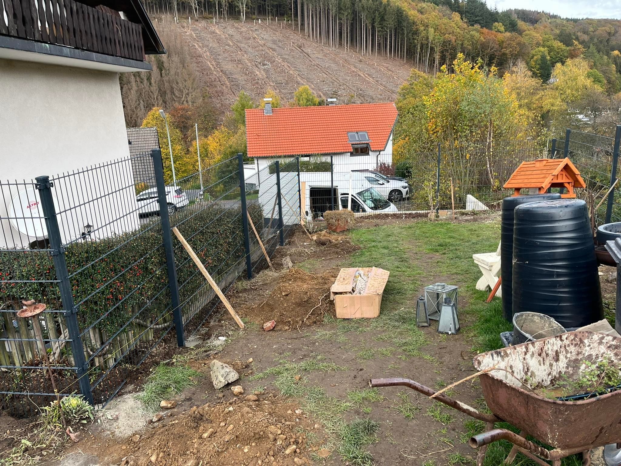 Montaż segmentów ogrodzenia panelowego na nierównym terenie, widoczne narzędzia, wykopana ziemia, tło z domem i lasem.