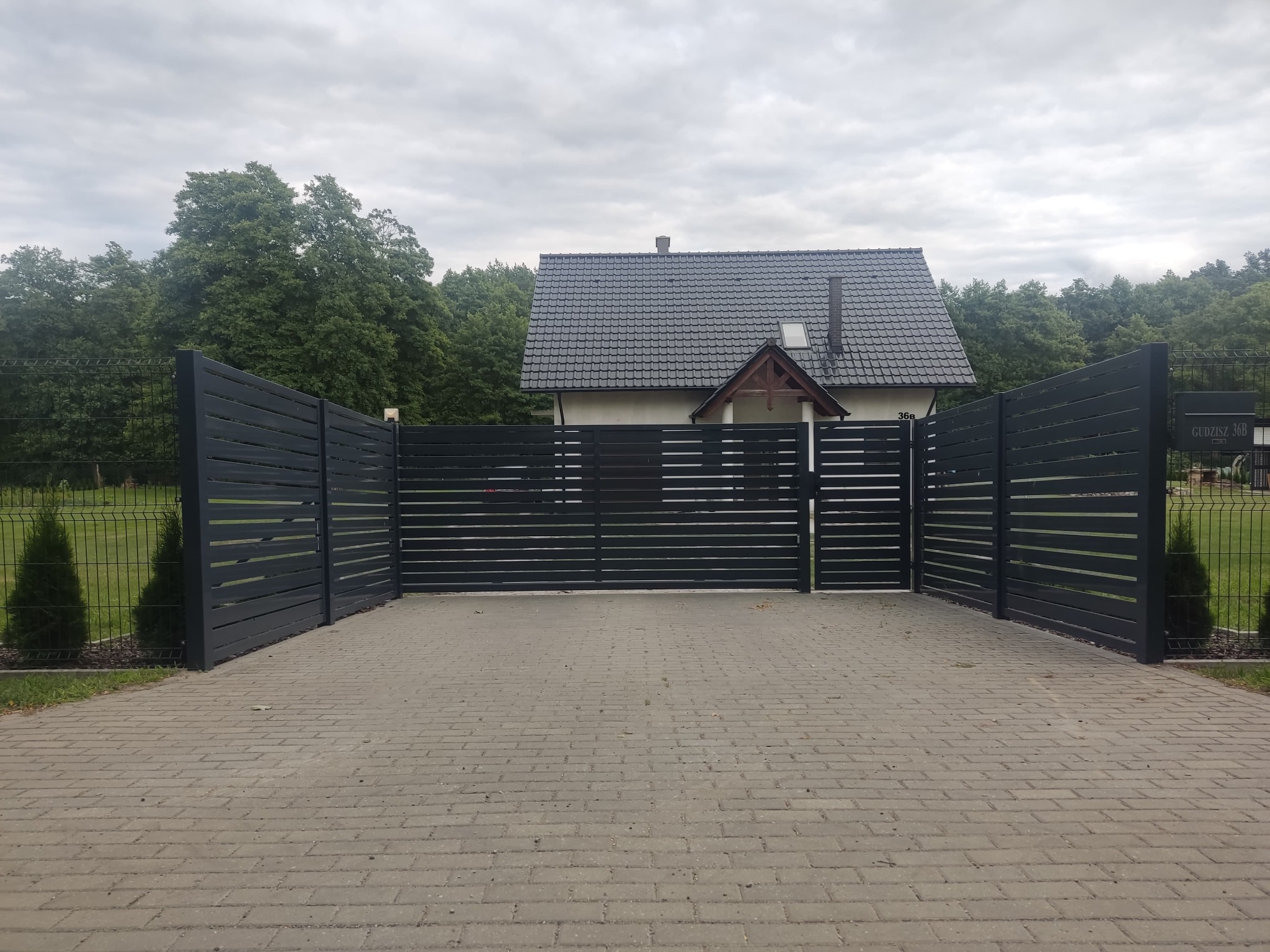 Nowoczesna, pozioma brama wjazdowa i ogrodzenie panelowe w kolorze grafitowym, prowadzące do domu z ciemną dachówką, widoczne z perspektywy podjazdu z kostki brukowej.