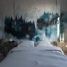 Sypialnia z łóżkiem, drewnianymi szafkami nocnymi, lampami wiszącymi i tapetą z motywem leśnym w odcieniach niebieskiego i szarego, z widocznym logotypem projektanta w prawym dolnym rogu.