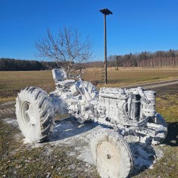Firma Usługowo- Handlowa Patryk Włoch - Traktor pokryty pianą czyszczącą na tle pola i lasu w słoneczny dzień. Widoczny maszt oświetleniowy i drzewo bez liści. Nietypowe ujęcie maszyny rolniczej po piaskowaniu.