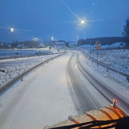 Firma Usługowo- Handlowa Patryk Włoch - Zimowy przejazd drogą ekspresową widziany z kabiny pojazdu odśnieżającego. Śnieg na drodze i poboczach, w tle oświetlone budynki i drzewa.
