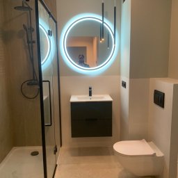 Zygnet Zyga Paweł - Nowoczesna łazienka z kabiną prysznicową typu walk-in z czarnymi profilami, okrągłym lustrem z podświetleniem LED, wiszącą szafką z umywalką i podwieszaną toaletą.