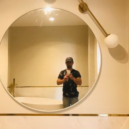 Zygnet Zyga Paweł - Okrągłe lustro w łazience z białymi płytkami i złotymi akcentami, odbicie mężczyzny robiącego zdjęcie telefonem.