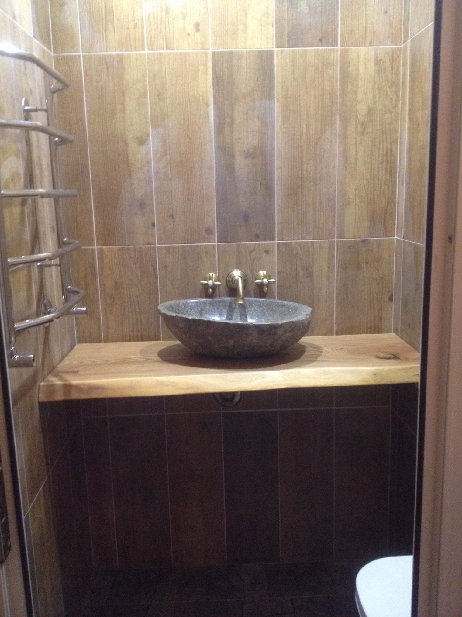 Kamienna umywalka w kształcie misy z mosiężną armaturą na blacie z naturalnego drewna w łazience wyłożonej płytkami imitującymi drewno.